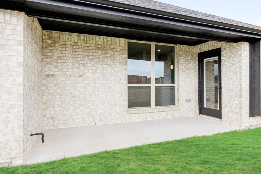 Covered back patio with concrete floor, light brick exterior, large window, and black door with green lawn.