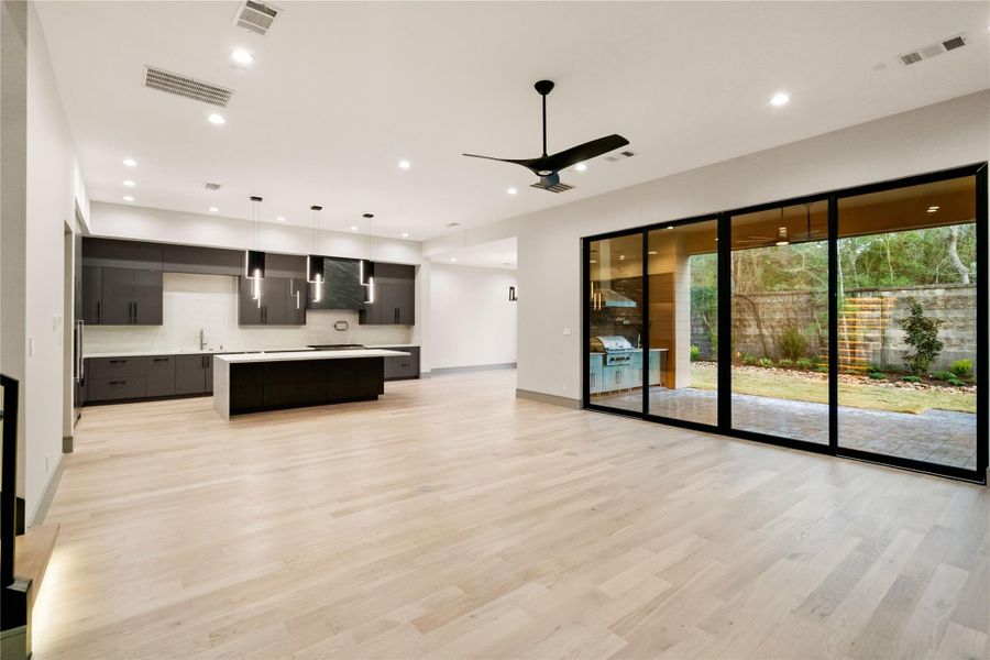This modern open-concept living space features a sleek kitchen with dark cabinetry, a large island, and pendant lighting. It has light wood flooring and expansive sliding glass doors leading to a patio and landscaped yard, offering ample natural light and outdoor views.