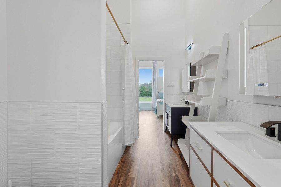 This photo showcases a bright and modern bathroom with sleek white fixtures, wooden flooring, and ample shelving. There's a bathtub with a shower curtain, a large mirror, and an open view leading to a bedroom with natural light.