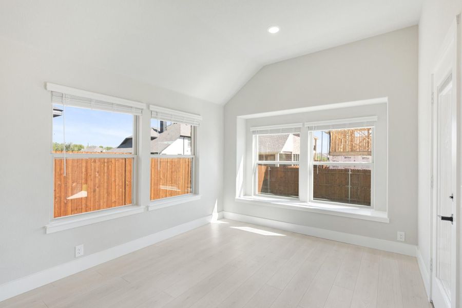 Empty bedroom with vaulted ceiling, light wood floors, and multiple windows with backyard views