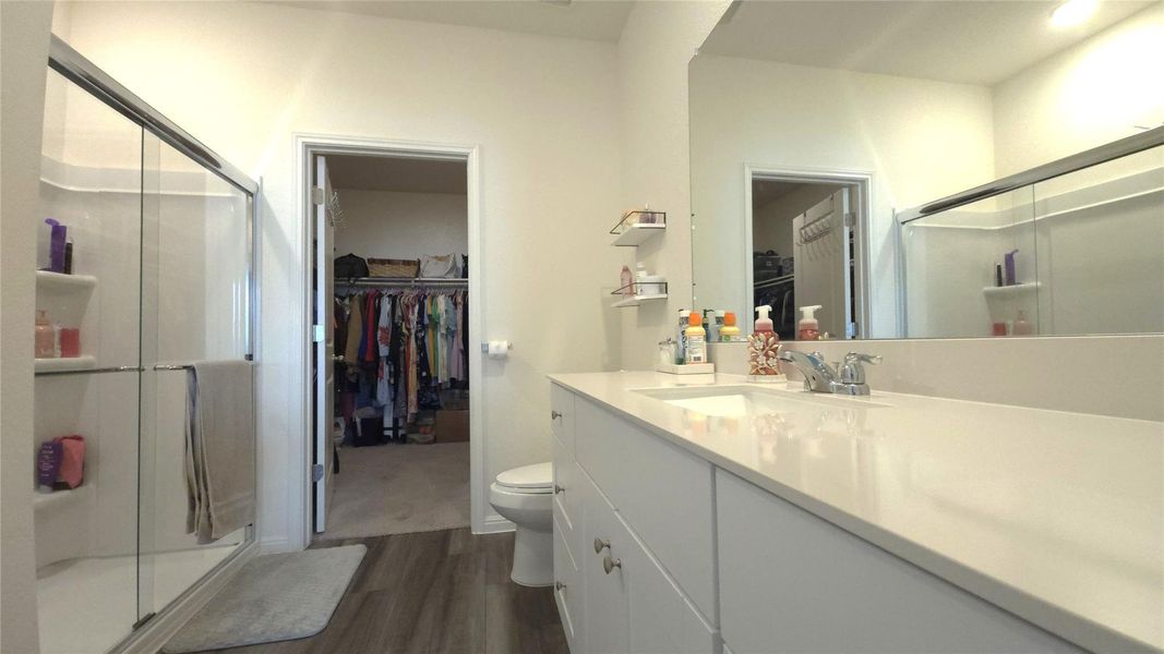 Bathroom featuring a spacious double vanity with a white countertop and undermount sinks, a glass-enclosed shower with built-in shelving, and wood-finish flooring