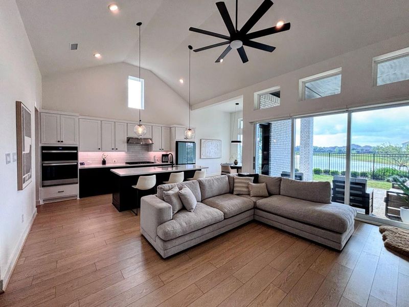 Spacious living area features modern design with high ceilings, a large ceiling fan, and ample natural light. The open layout connects a sleek kitchen with white cabinetry and a central island to a cozy living room. Expansive windows offer views of a serene backyard and lake, enhancing the inviting ambiance.