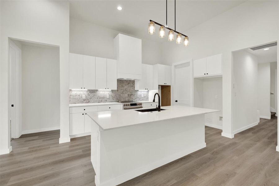 Kitchen featuring hanging light fixtures, a towering ceiling, white cabinets, a center island with sink, and light wood-type flooring