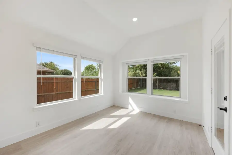 Empty bedroom with vaulted ceiling, light wood floors, and four windows overlooking a fenced backyard