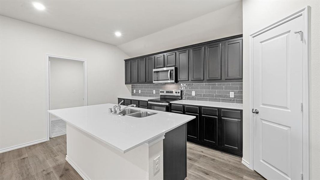 Kitchen featuring a central island with a double basin sink, dark cabinetry, light countertops, and a gray subway tile backsplash