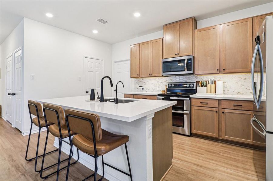 Kitchen with decorative backsplash, appliances with stainless steel finishes, a kitchen bar, a center island with sink, and light wood-style floors Kitchen with decorative backsplash, appliances with stainless steel finishes, a kitchen bar, a center island with sink, and light wood-style floors
