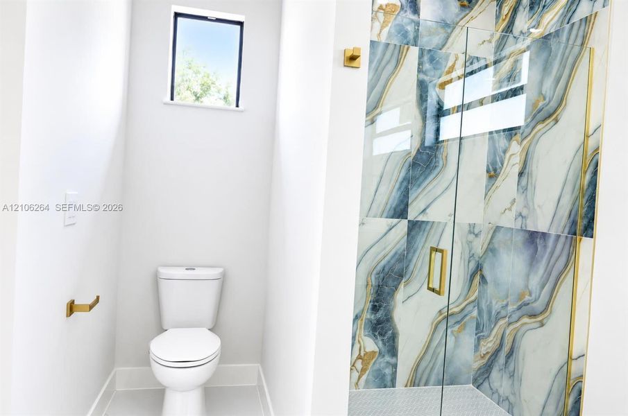 Shower enclosure featuring large-format decorative tile with gold-tone metal trim and hardware, clear glass paneling, and a high-efficiency toilet
