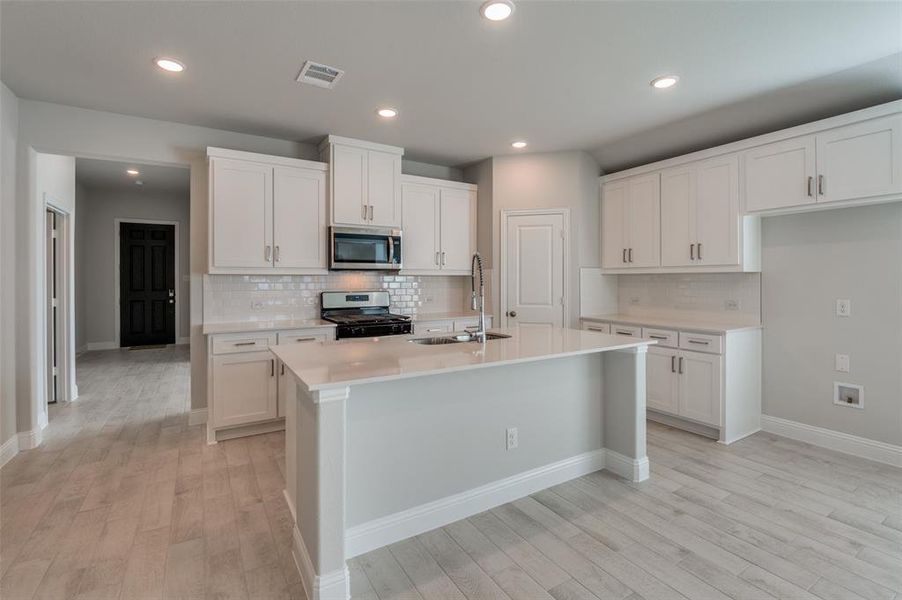 Kitchen featuring an island with sink, white shaker cabinetry, stainless steel appliances, subway tile backsplash, and light wood-finish flooring