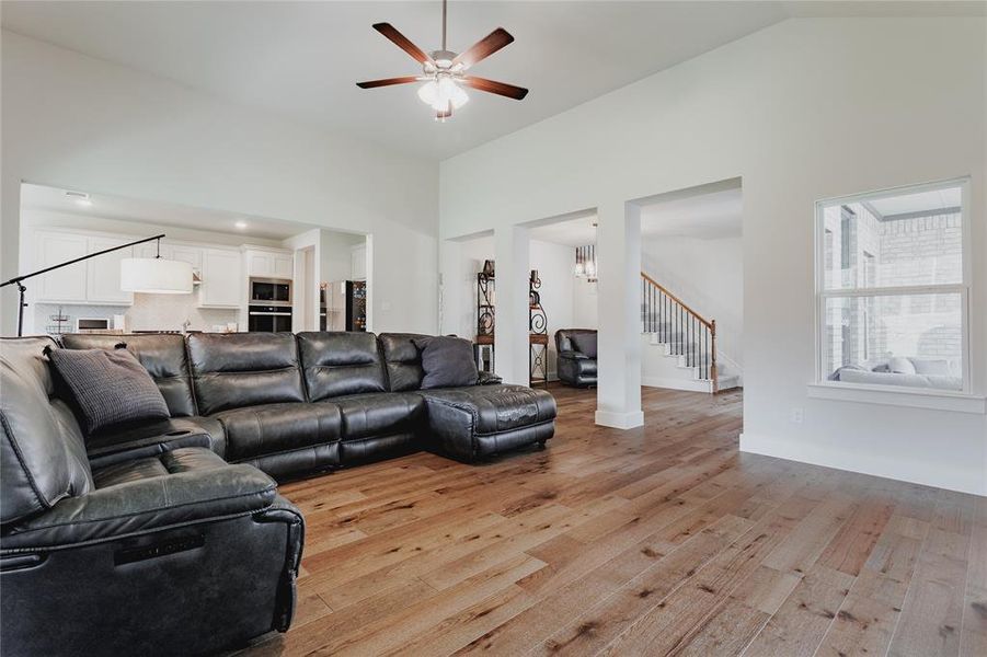Living area featuring high vaulted ceilings, wood-finish flooring, a ceiling fan with integrated lighting, and a clear sightline to the kitchen with white cabinetry and built-in appliances