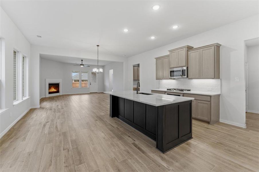 Spacious kitchen featuring a large island with a light-toned countertop, light-toned cabinetry, and stainless steel appliances, opening to a living area with a fireplace and wood-finish flooring