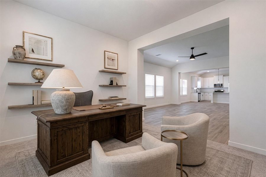 This inviting office space offers a clear view back toward the entry and adjacent rooms, showcasing the home’s thoughtful, open layout. A modern ceiling fan adds comfort and style, while the combination of tile flooring and soft carpeted transitions defines each space. Natural light and clean finishes create a bright, welcoming atmosphere throughout. This photo is virtually staged.