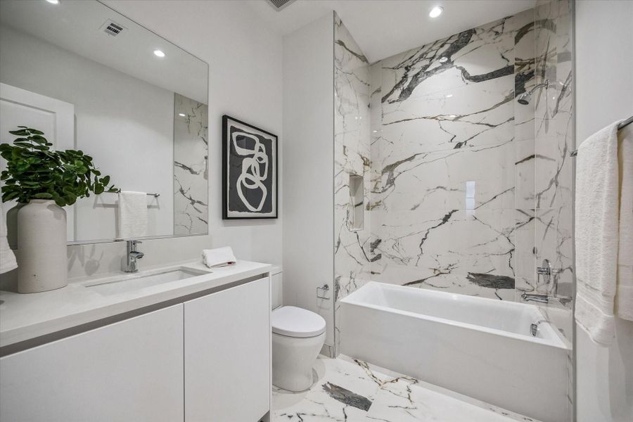 This secondary bath features a clean, modern design with a tub/shower combination wrapped in striking marble, complemented by a sleek vanity & contemporary finishes.