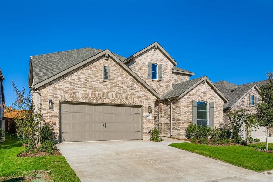 French country inspired facade with a shingled roof, brick siding, driveway, a front yard, and an attached garage