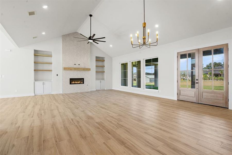 Unfurnished living room featuring built in shelves, light wood-style flooring, a chandelier, a fireplace, and high vaulted ceiling Unfurnished living room featuring built in shelves, light wood-style flooring, a chandelier, a fireplace, and high vaulted ceiling