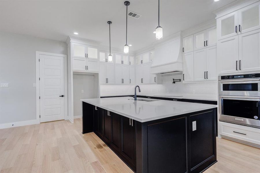 Kitchen featuring a large central island with a white countertop and dark cabinetry, white perimeter cabinetry with glass-front upper cabinets, stainless steel double ovens, and wood-finish flooring