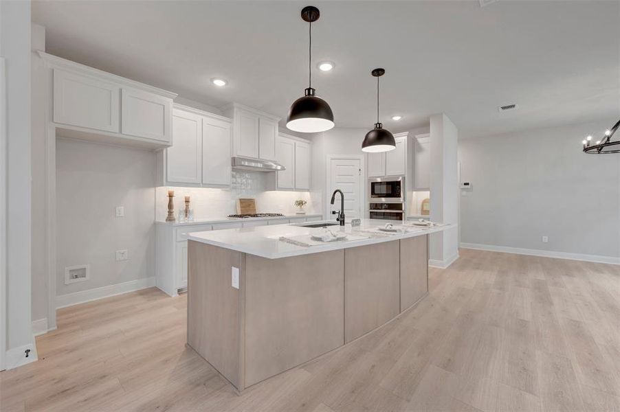 Open-concept kitchen featuring an oversized island with seating, white shaker cabinetry, stainless steel appliances including a built-in oven and microwave, and wood-finish flooring