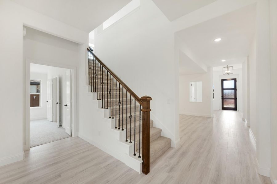 Bright entryway with carpeted staircase, wood railing, iron balusters, and light hardwood floors leading to front door.