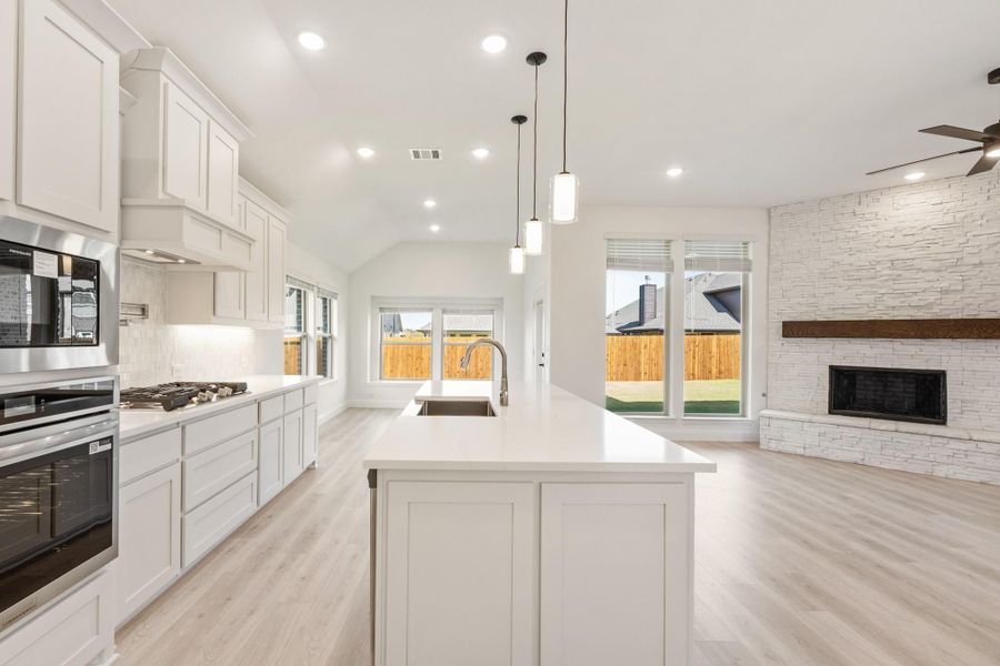 Open-concept kitchen with white cabinets, large island, and stone fireplace visible in adjoining living area Open-concept kitchen with white cabinets, large island, and stone fireplace visible in adjoining living area