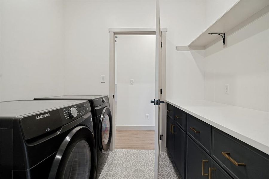 Laundry area featuring patterned tile flooring, dark cabinetry with brass hardware, a white countertop, and an upper wall shelf