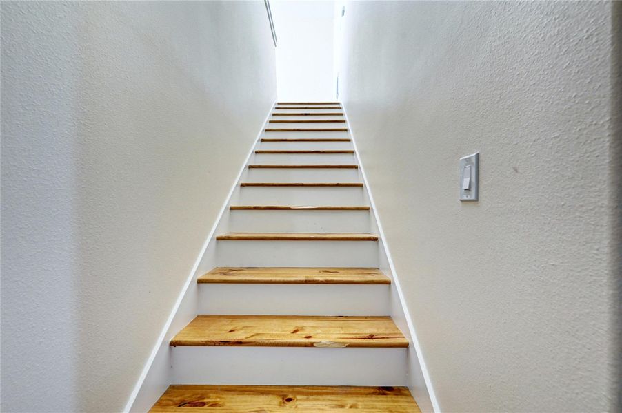 The photo shows a staircase with light wood steps and white risers, leading upward between two plain walls, creating a clean, modern look.