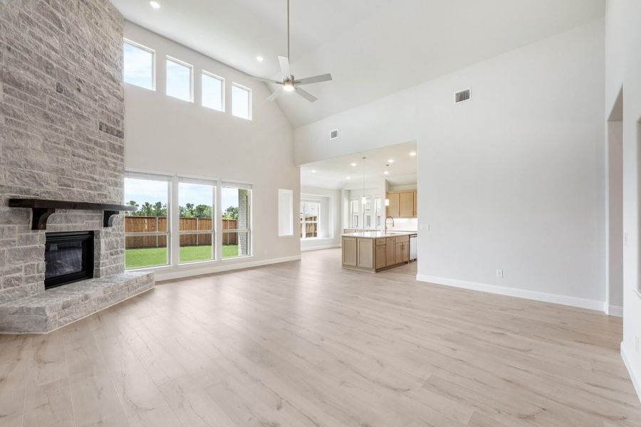Open living room with stone fireplace, high vaulted ceiling, and light wood floors open to kitchen