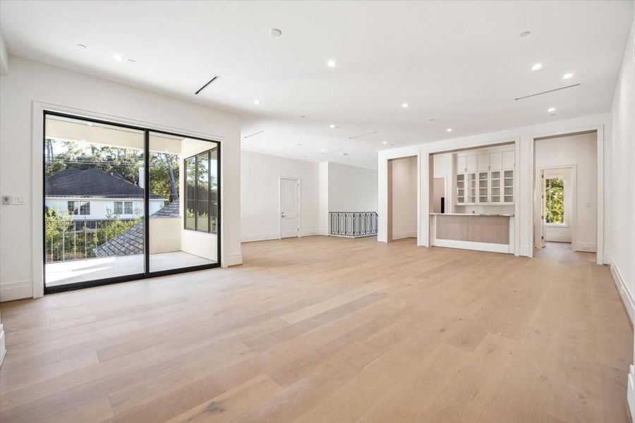 An expansive game room with engineered white oak flooring, recessed lighting, and a sliding door that opens to the covered balcony. A built-in refreshment area with custom cabinetry enhances the space, making it ideal for entertaining, lounging, or serving as a versatile second-floor living retreat.