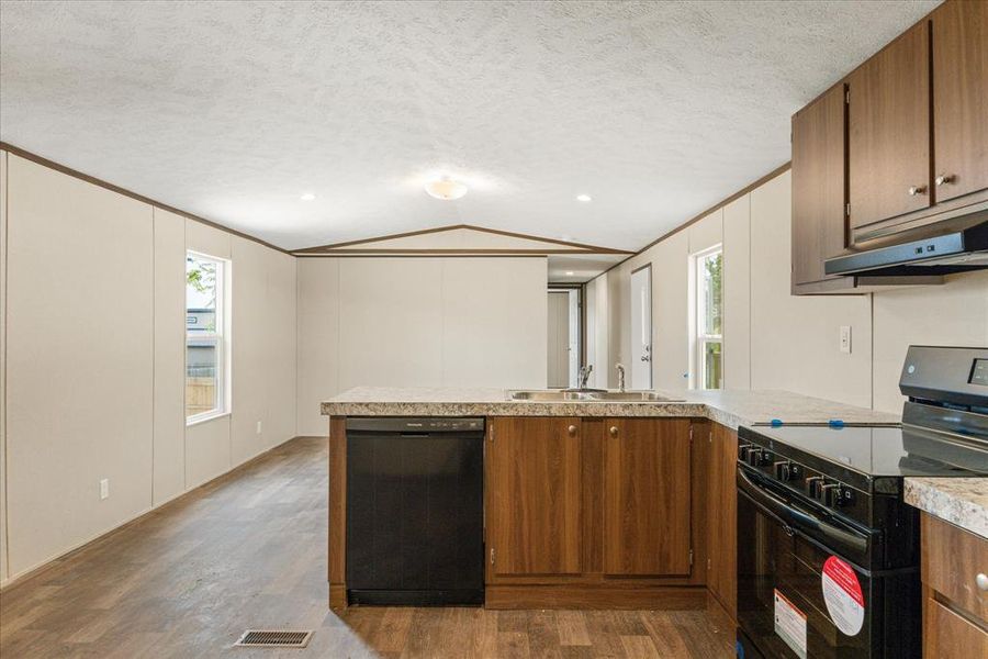 Kitchen featuring wood-finish cabinetry, light-toned countertops, a stainless steel double basin sink, and integrated black appliances including a dishwasher and range with an overhead vent