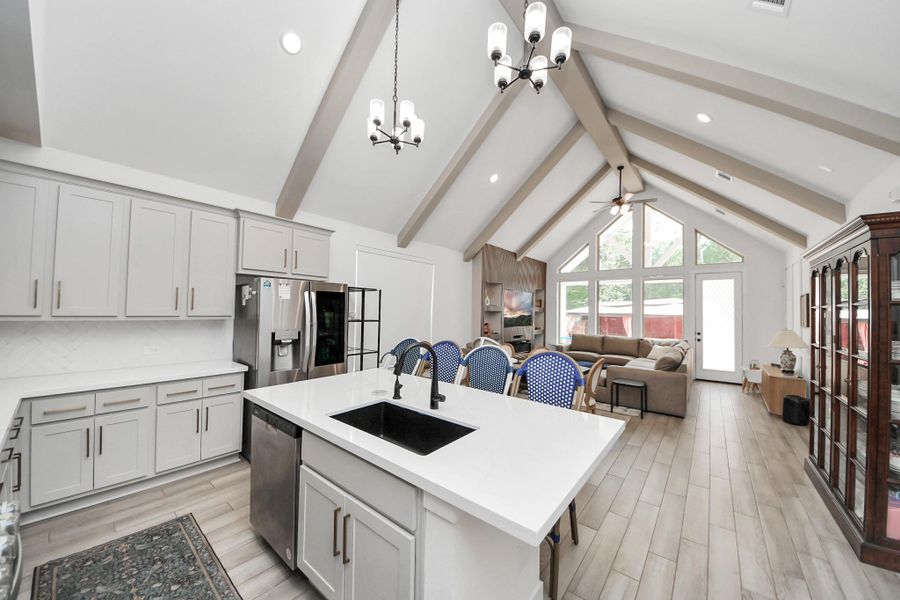 This open-concept kitchen and living area features vaulted ceilings with exposed beams, modern light fixtures, and ample natural light from large windows. The kitchen includes sleek white cabinets, stainless steel appliances, and a central island with a sink. The living space is cozy, with a comfortable seating area and access to the backyard. This open-concept kitchen and living area features vaulted ceilings with exposed beams, modern light fixtures, and ample natural light from large windows. The kitchen includes sleek white cabinets, stainless steel appliances, and a central island with a sink. The living space is cozy, with a comfortable seating area and access to the backyard.