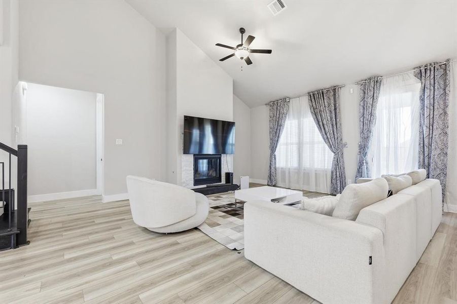 Living room with high vaulted ceiling, a glass covered fireplace, light wood-style flooring, ceiling fan, and stairs