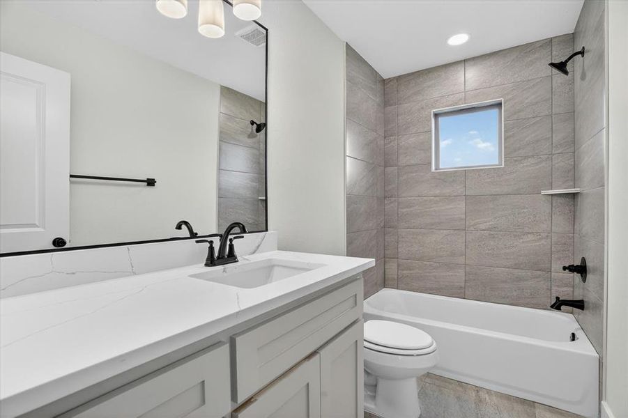 Bathroom featuring a vanity with a white countertop and undermount sink, a large mirror with a black frame, light gray shaker-style cabinetry, and a bathtub with gray tile surround and matte black fixtures