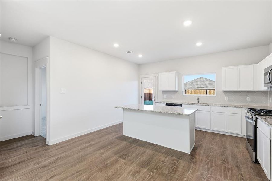 Spacious kitchen featuring an island, white cabinetry, integrated stainless steel appliances, speckled stone countertops, and wood-finish flooring Spacious kitchen featuring an island, white cabinetry, integrated stainless steel appliances, speckled stone countertops, and wood-finish flooring