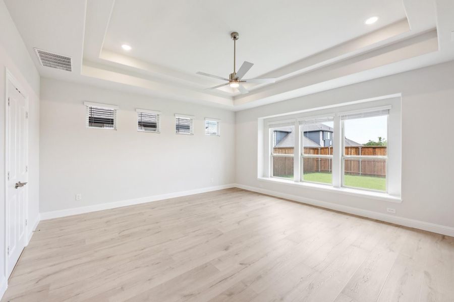 Empty bedroom with tray ceiling, ceiling fan, light wood floors, and large backyard-facing window