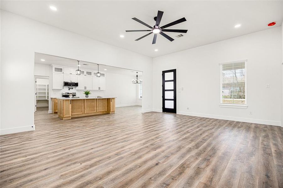 Bright, open-concept living space with modern finishes. Features hardwood floors, a large ceiling fan, and a spacious kitchen with an island and white cabinetry. Natural light fills the room through multiple windows, enhancing the home's airy feel.