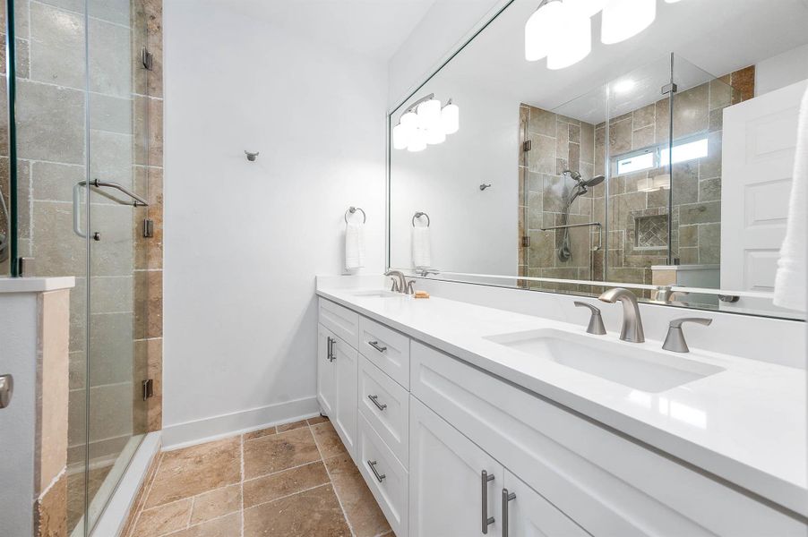 The primary bathroom offers a spacious and modern design featuring dual sinks, expansive counter space, and sleek white cabinetry. A beautifully tiled glass-enclosed shower adds a touch of luxury while creating a clean and elegant atmosphere.