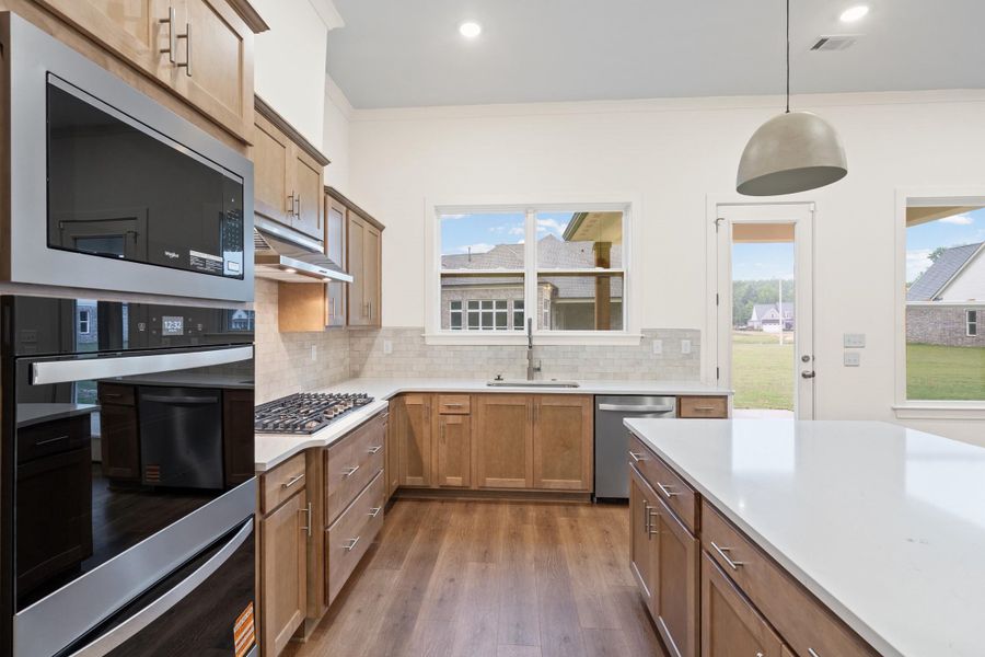 Open-concept kitchen featuring wood-finish flooring, light wood cabinetry, white countertops, and stainless steel appliances including a wall oven, microwave, and gas cooktop Open-concept kitchen featuring wood-finish flooring, light wood cabinetry, white countertops, and stainless steel appliances including a wall oven, microwave, and gas cooktop