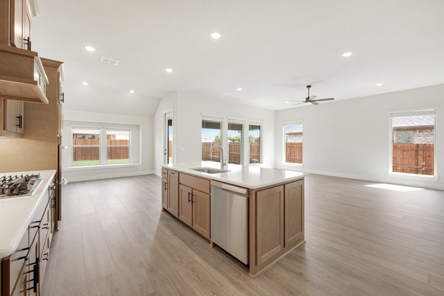 Open-concept kitchen with center island, sink, stainless dishwasher, wood cabinets, and light hardwood floors flowing into living area