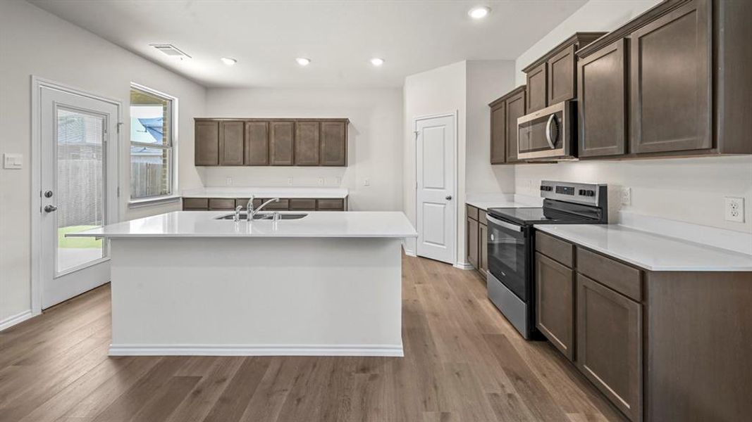 Open-concept kitchen featuring a large central island with an undermount sink, sleek white countertops, dark wood-finish cabinetry, stainless steel appliances, and wood-finish flooring