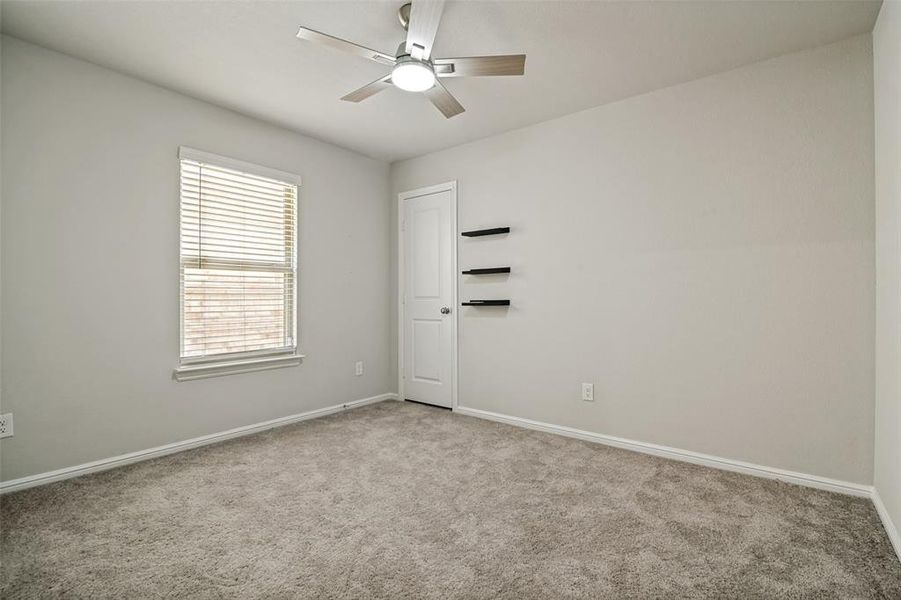 Room featuring light gray walls, gray carpet flooring, a window with blinds, a ceiling fan with integrated lighting, and a white door with black floating shelves Room featuring light gray walls, gray carpet flooring, a window with blinds, a ceiling fan with integrated lighting, and a white door with black floating shelves