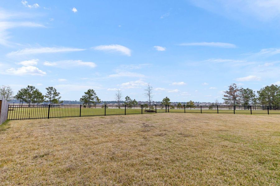 Spacious backyard with a well-maintained lawn, enclosed by a black metal fence, offering clear views of open fields and distant trees under a clear blue sky. Ideal for outdoor activities and relaxation. Spacious backyard with a well-maintained lawn, enclosed by a black metal fence, offering clear views of open fields and distant trees under a clear blue sky. Ideal for outdoor activities and relaxation.