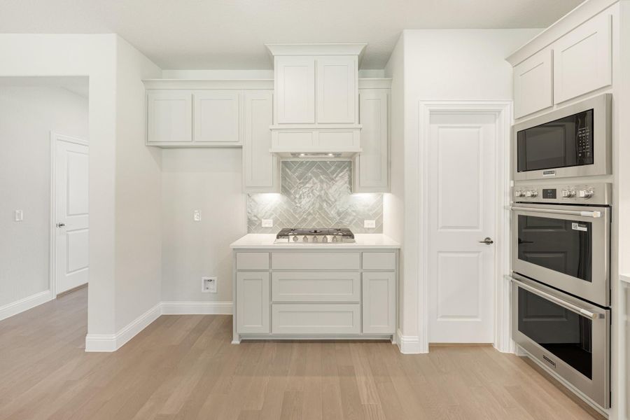 White kitchen with gas cooktop, herringbone tile backsplash, built-in double oven, and light wood floors