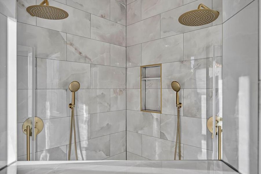 Dual shower system featuring gold-tone fixtures, rain showerheads, handheld sprayers, and marble-look tiled walls with a recessed niche Dual shower system featuring gold-tone fixtures, rain showerheads, handheld sprayers, and marble-look tiled walls with a recessed niche