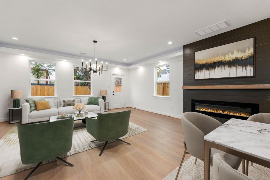 Spacious open-concept living area featuring warm wood flooring, recessed lighting, and abundant natural light. The focal point is a sleek linear fireplace with a modern accent wall and floating mantle. A tray ceiling with contemporary chandelier adds architectural interest, while the seamless flow into the dining and kitchen areas makes this space ideal for everyday living and entertaining.