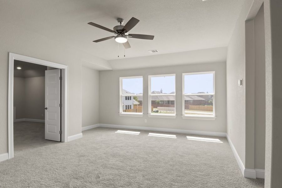 The primary bedroom with three white-framed windows, tan carpet, a brown ceiling fan and a open door.