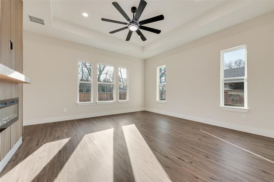 Primary Bedroom featuring a glass covered fireplace, a tray ceiling, light wood finished floors, ceiling fan, and recessed lighting