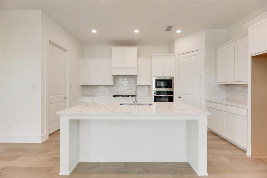 Kitchen with light stone countertops, white cabinets, an island with sink, stainless steel oven, and light wood-style floors Kitchen with light stone countertops, white cabinets, an island with sink, stainless steel oven, and light wood-style floors