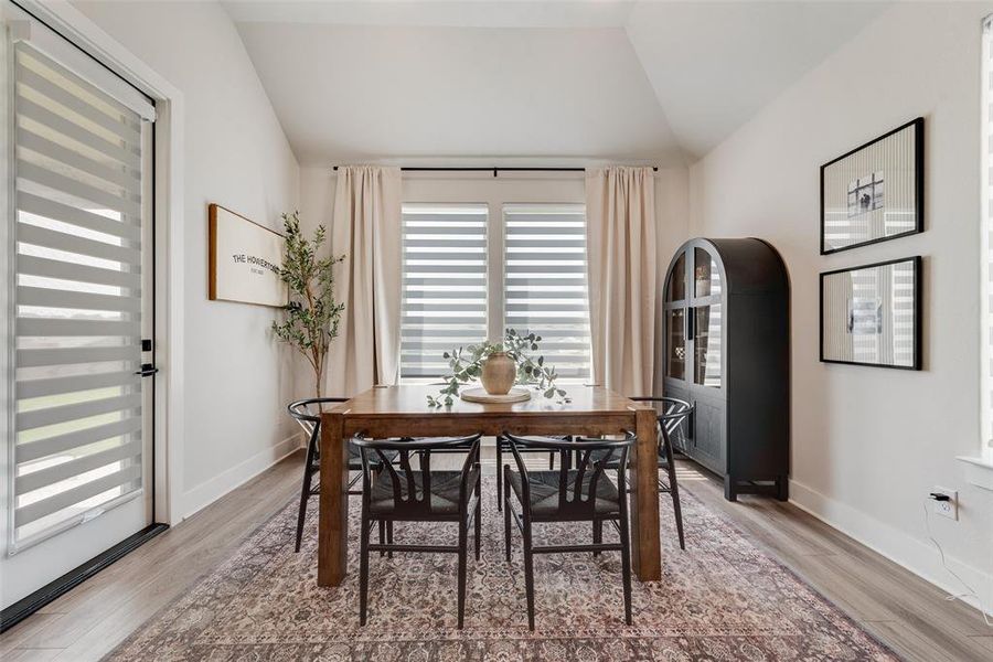 Vaulted ceiling dining area featuring light wood-finish flooring, a contemporary black arched display cabinet, and a large window with privacy blinds