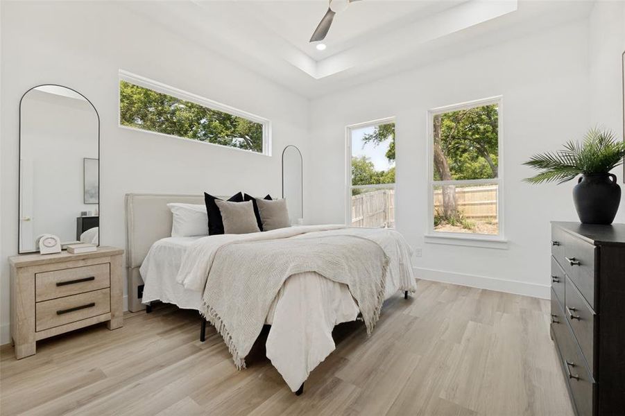 Bright sleeping area featuring wood-finish flooring, a tray ceiling with a ceiling fan, and multiple windows Bright sleeping area featuring wood-finish flooring, a tray ceiling with a ceiling fan, and multiple windows