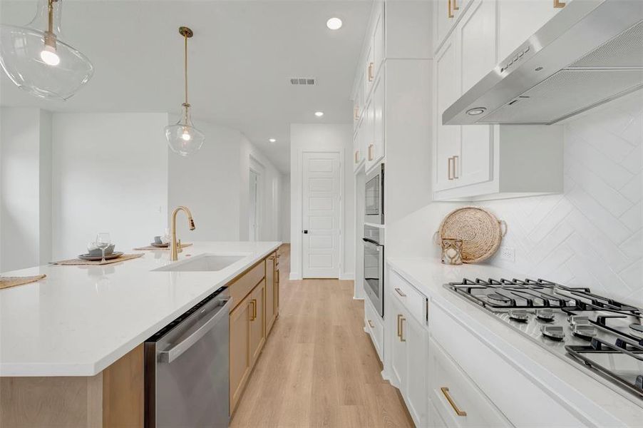 Kitchen featuring white cabinetry, light wood-finish flooring, a central island with an undermount sink and brass-finish faucet, and stainless steel appliances including a gas range and built-in ovens
