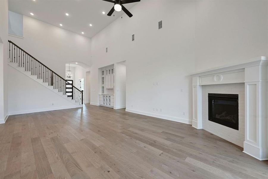 Unfurnished living room featuring stairs, light wood finished floors, a towering ceiling, ceiling fan, and a glass covered fireplace Unfurnished living room featuring stairs, light wood finished floors, a towering ceiling, ceiling fan, and a glass covered fireplace