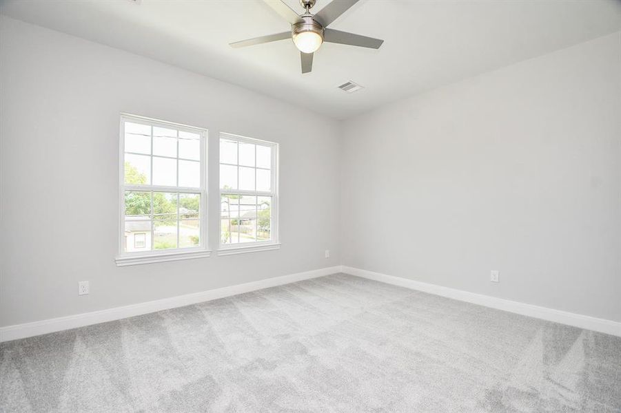 This is a bright, airy bedroom with neutral walls, plush carpeting, and a ceiling fan. There are two large windows providing natural light and a view of the outside.